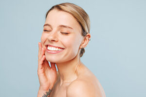 Close up happy half naked topless young woman 20s with nude make up close eyes with toothy smile touch face isolated on pastel blue color background.